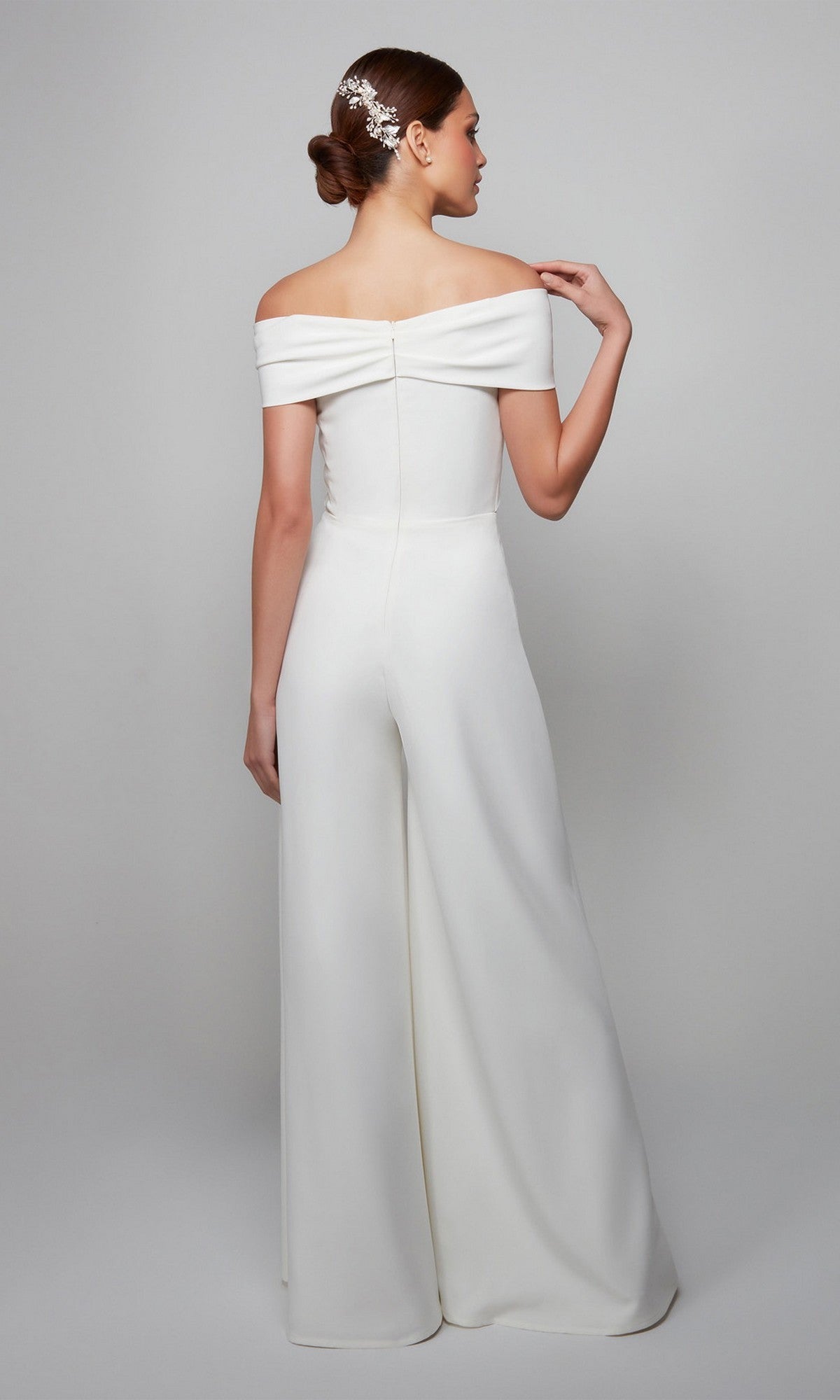 Woman in an Alyce 70013 long formal off-the-shoulder jumpsuit with wide legs, seen from the back; her hair is styled in a low bun with a decorative hairpiece.